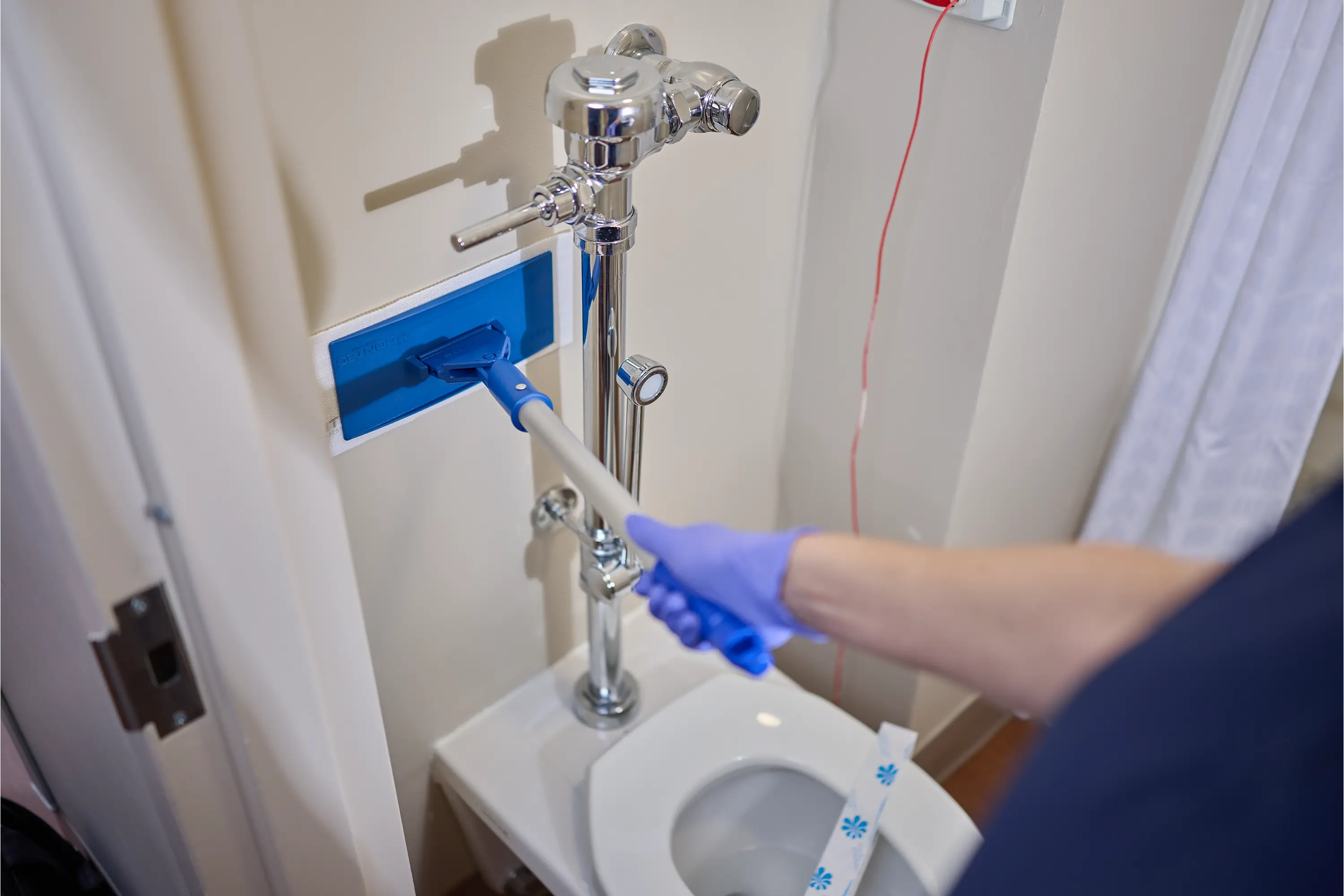 Environmental services worker using a Tight Quarters mop to clean a narrow space beside a toilet in a healthcare facility restroom.