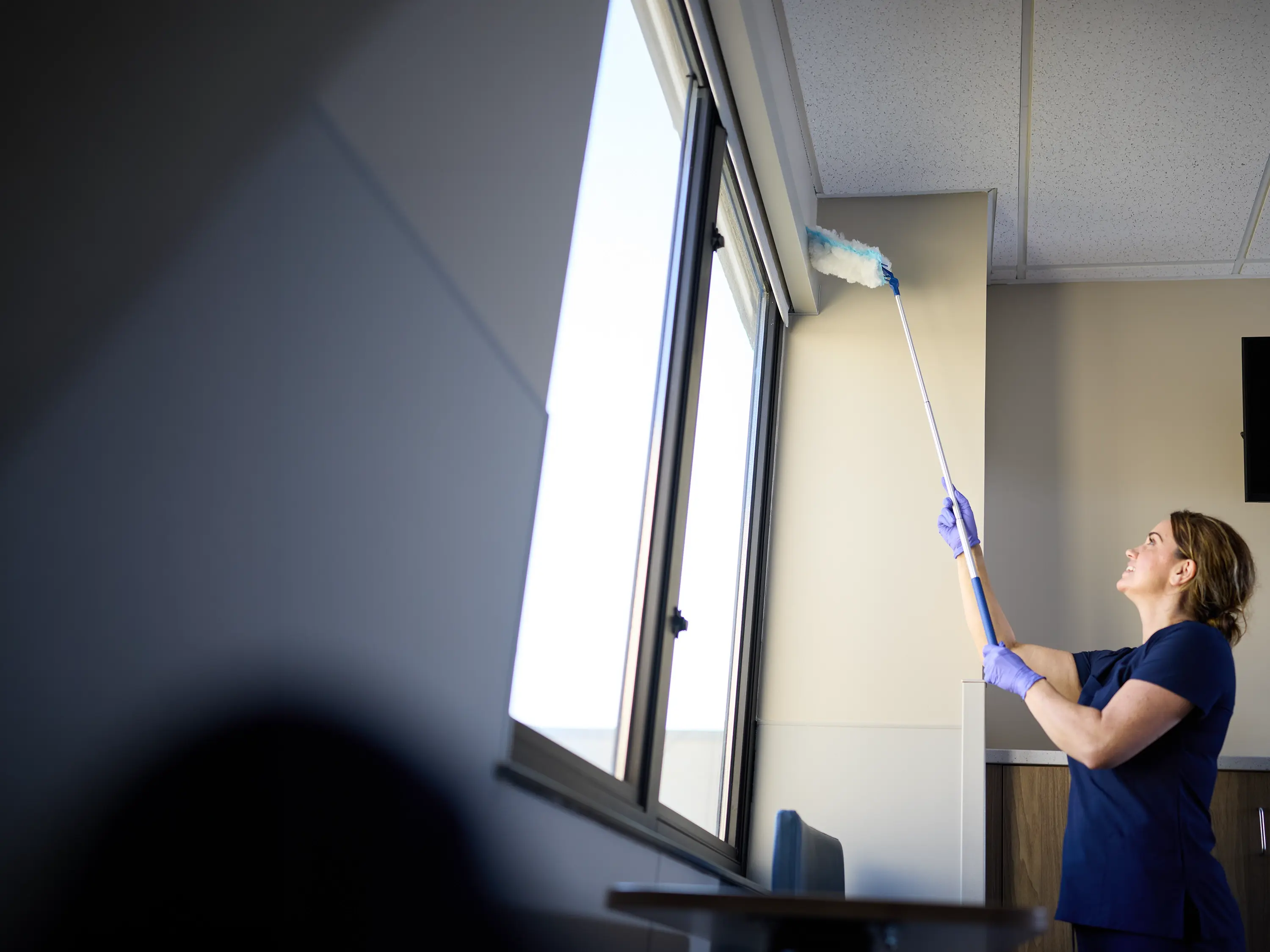 Environmental services worker using an extendable duster to clean a high window frame in a healthcare facility.