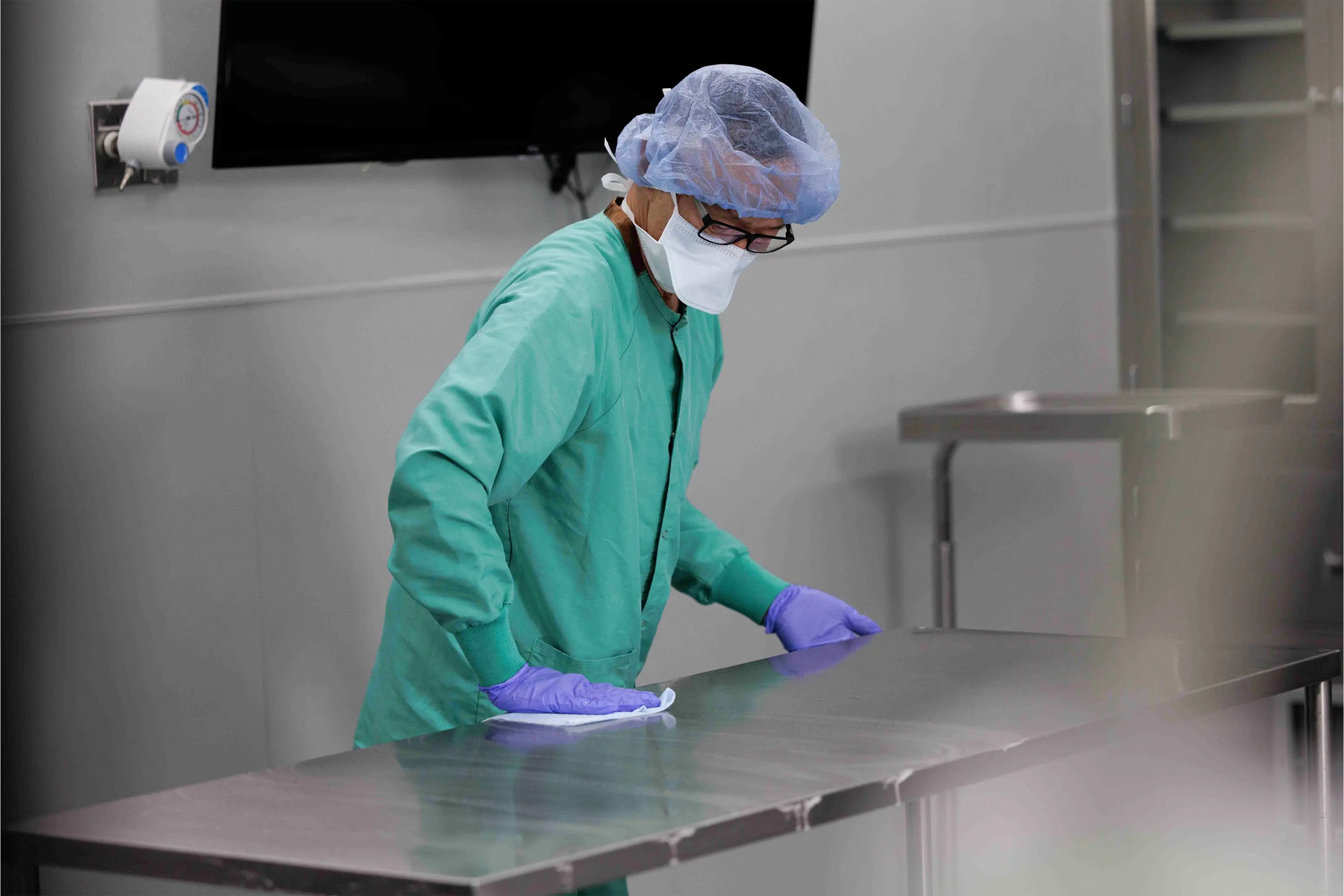 Healthcare worker in surgical attire wiping a stainless steel work table in an operating room.