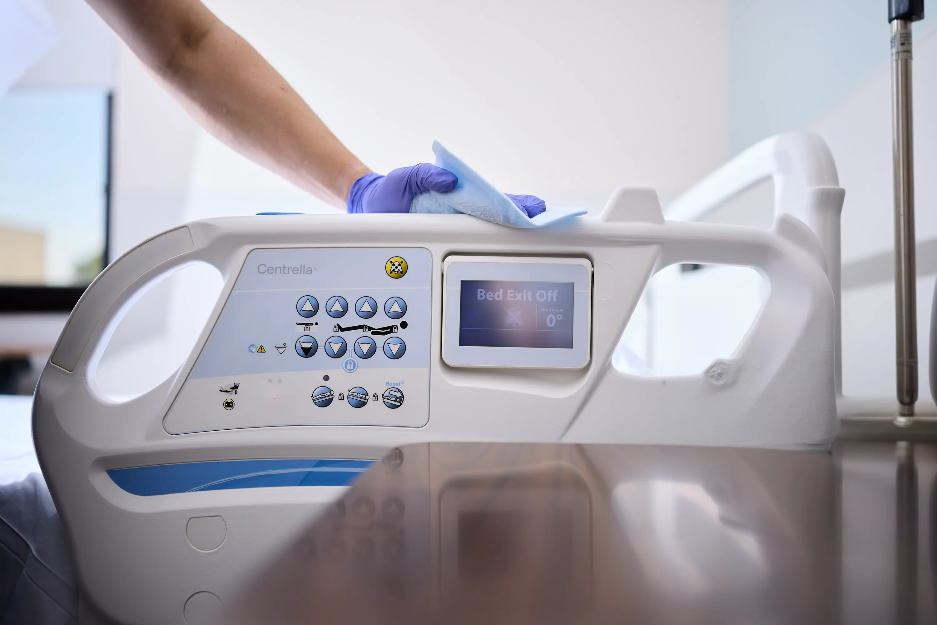 Purple-gloved worker wiping the side rail and controls of a hospital bed in a patient care environment.