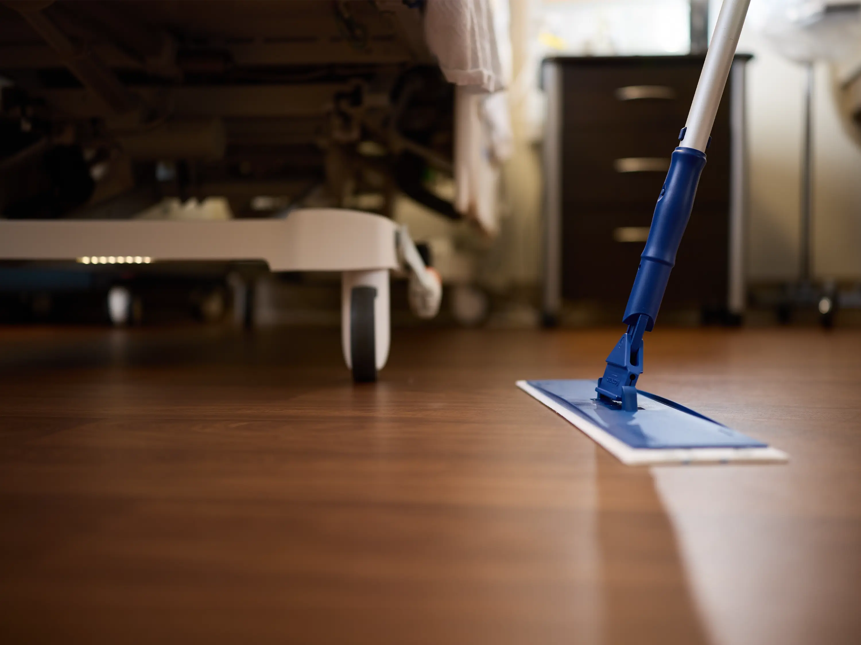 Low-angle view of a mop cleaning the floor beside a patient bed in a healthcare environment.