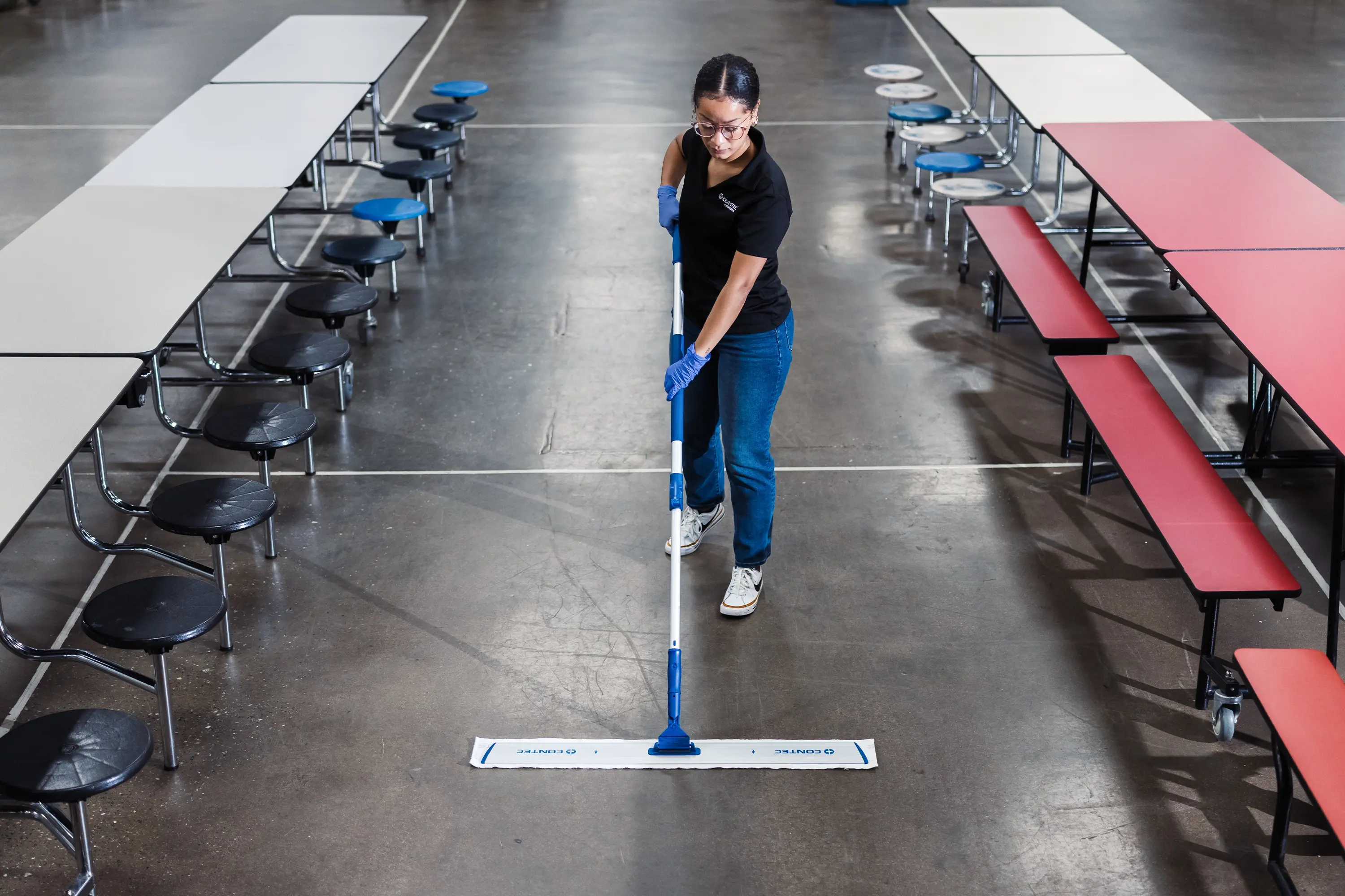 Worker mopping the floor between rows of cafeteria tables in a large dining area.