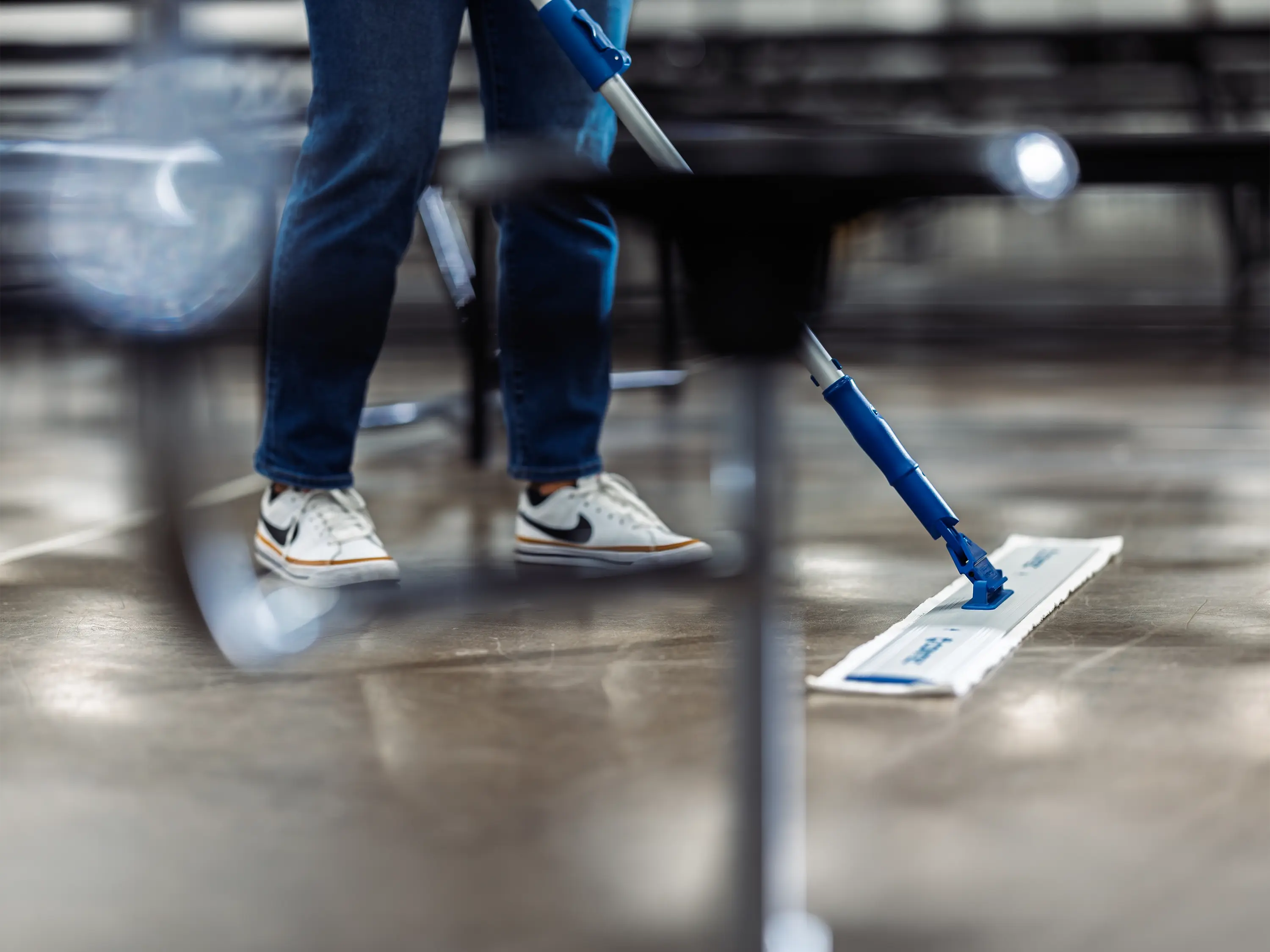 Worker mopping a hard-surface floor beneath tables in a large public venue.